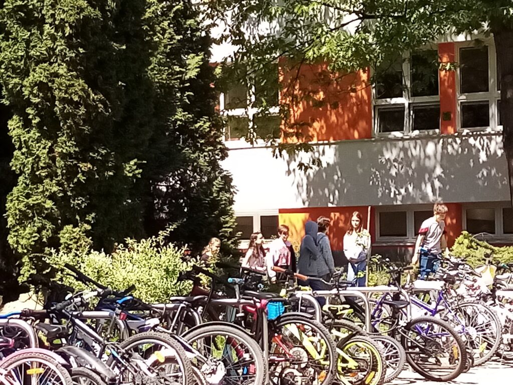 zdjęcia uczniów klasy 7d na terenie szkolnego ogródka; widoczne głównie postaci w oddali, drzewa i krzewy. Na 1 ze zdjęć widać także rowery i hulajnogi, na których uczniowie przyjechali do szkoły. 